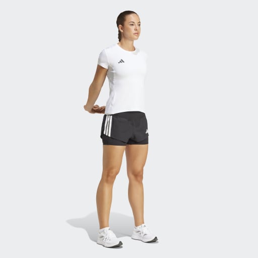 En person står på en almindelig lysegrå baggrund iført en hvid atletisk t-shirt, Adidas Adizero 2I1 Shorts Dame i sort for optimal løbeydelse og hvide atletiske sko. Armene er bag ryggen, mens de kigger lidt til siden.