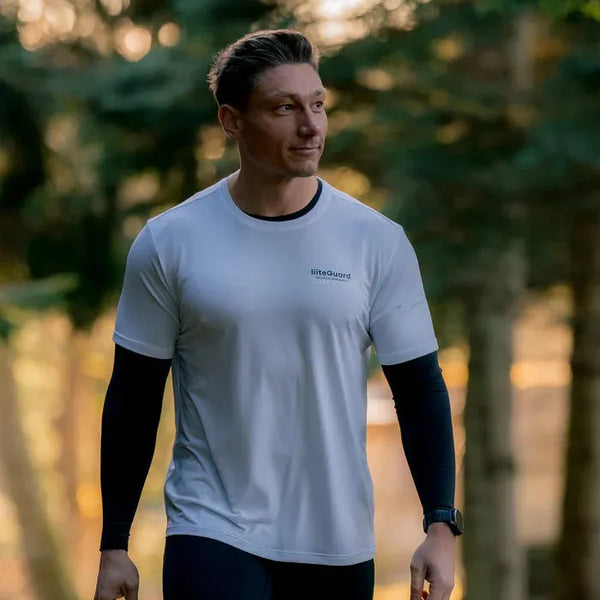 En mand med kort brunt hår og et armbåndsur går udendørs foran grønne træer iført en LiiteGuard Ground-Tech T-shirt, Herre, White over en sort langærmet skjorte. Sollyset filtreres gennem den skovklædte baggrund.