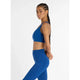 En kvinde med langt flettet hår bærer New Balance Define Train Bra til kvinder i Galaxy Blue og matchende leggings, mens hun står sidelæns med hænderne foldet bag ryggen mod en almindelig hvid baggrund.