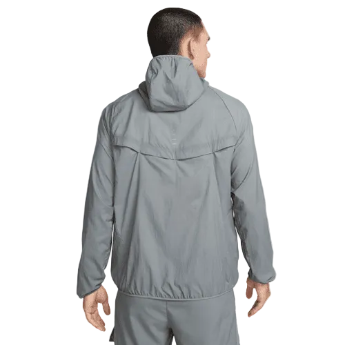 En mand kigger væk fra kameraet i en Nike Repel UV Running Jacket, Herre, Smoke Grey/Smoke Grey/Reflective Silv - en ideel løbejakke herre med UV-beskyttelse - sammen med matchende grå bukser på en almindelig hvid baggrund.