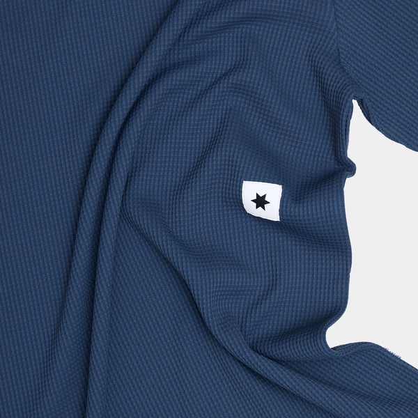 Et nærbillede af Saysky Motion Waffle Long Sleeve, Herre, Blue viser det strukturerede stof og et lille hvidt mærke med en sort stjerne på en lys baggrund.