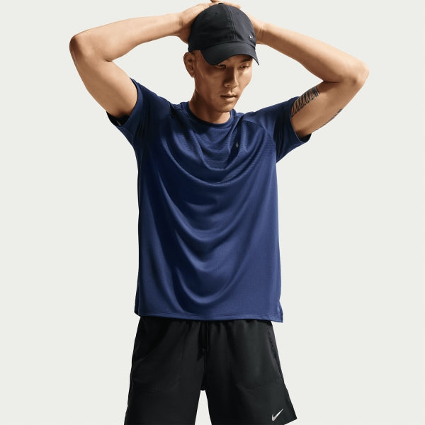 En mand i mørkeblå sportstee, Nike Stride 7" Brief-Lined Running Shorts (Black/Reflective Silver) og en sort kasket står med hænderne bag hovedet. Synlige Nike-logoer. Baggrunden er ensfarvet og lys.