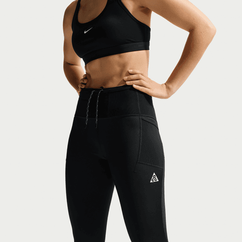 En kvinde bærer en sort sports-bh og Nike ACG High-Waisted 7/8 Running Tights, Dame, i Black/Summit White, med hænderne på hofterne. Nike- og ACG-logoer er synlige på tightsene. Vist fra skuldre til lår.