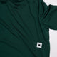En mørkegrøn Saysky Logo Pace Long Sleeve til mænd ligger fladt med et hvidt, rektangulært mærke med en sort stjerne syet i bunden af siden.