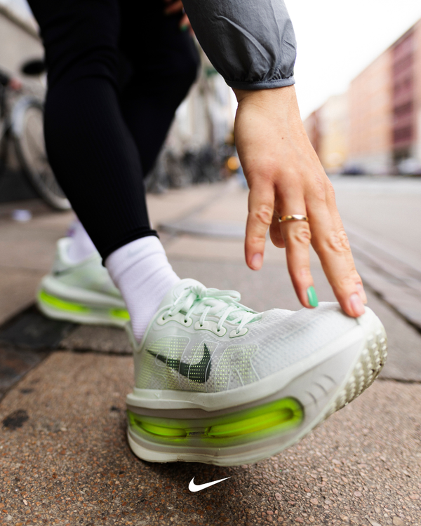 En person iført hvide Nike-sneakers med neongrønne detaljer bøjer sig ned for at røre ved tæerne på et fortov i byen. Kun underbenene og den ene hånd er synlige. Nike-logoet er nederst i midten af billedet.