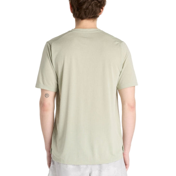 En person vender ansigtet væk fra kameraet og er iført en New Balance Athletics T-shirt til mænd i Garter Snake Heather og lyse shorts. En lille cirkulær tatovering er synlig på venstre overarm mod en almindelig hvid baggrund.