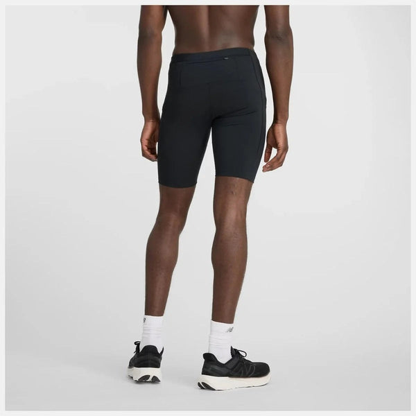 En person kigger væk fra kameraet iført New Balance NB Sleek Pocket Half Tight 9" Herre i sort sammen med hvide strømper og sorte sneakers på en almindelig lys baggrund.