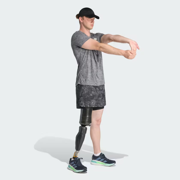 En person med en protese på højre ben, i sportstøj og kasket, strækker sig frem på en lys baggrund og viser Adidas Otr Sd 2In1 M herre løbeshorts i grefou/sort til stilfuld træning.