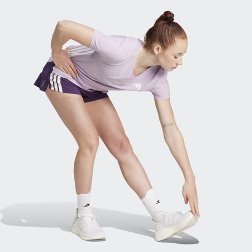 En kvinde iført Adidas Adizero Gel Dame Aurplu strækker sig frem og rører ved sin venstre fod med sin højre hånd; hun er klædt i en lyslilla t-shirt, lilla shorts, hvide sokker og sneakers på en ensfarvet baggrund.