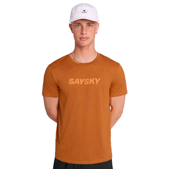 En mand iført Saysky Logo Pace T-shirt, Herre, i orange, en hvid kasket og sorte shorts står på en almindelig lys baggrund med hænderne bag ryggen.