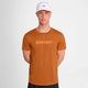 En mand iført Saysky Logo Pace T-shirt, Herre, i orange, en hvid kasket og sorte shorts står på en almindelig lys baggrund med hænderne bag ryggen.