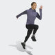 En person i lilla Adidas Otr Bin Hz Dame Previo træningstrøje, sorte leggings og sorte sneakers er fanget midt i et spring mod en lysegrå baggrund.