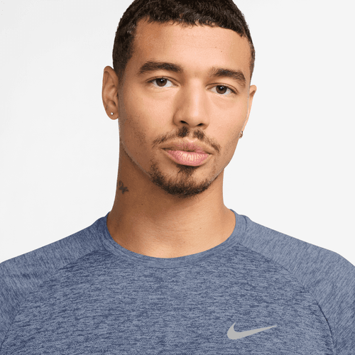 En mand med kort krøllet hår, let overskæg og fipskæg er iført en Nike Dri-FIT ADV kortærmet løbetrøje i World Indigo/Htr/Reflective Silv. Han har små øreringe, en lille halstatovering og står på en almindelig hvid baggrund.