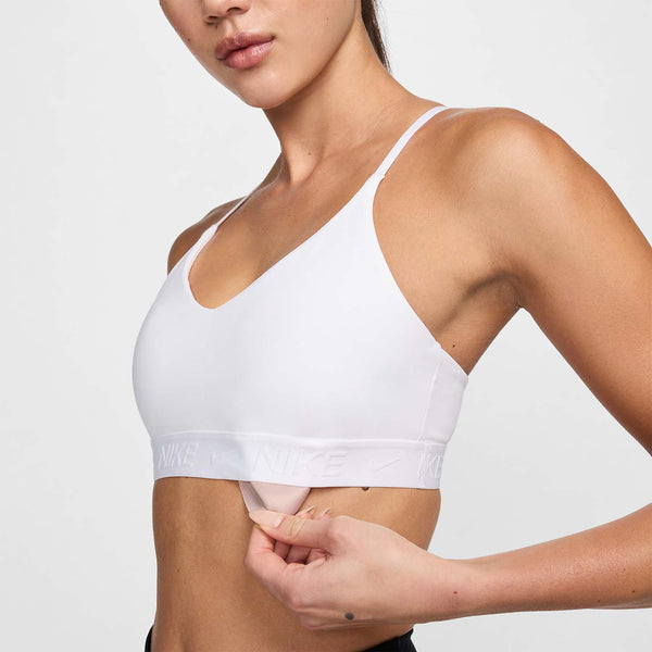 En kvinde bærer en hvid/stenlilla Nike Padded Adjustable Sports Bra og trækker den nederste kant op for at afsløre en inderlomme. Det beskårne billede viser hendes overkrop og en del af hendes ansigt.