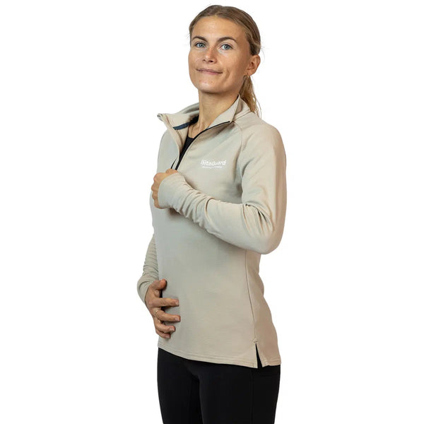 En kvinde smiler på en hvid baggrund iført Liiteguard Ground-Tech 1/4 Zip, Dame, Beige. Den svedtransporterende langærmede top går sammen med sorte bukser; hun kigger opad med en hånd på brystet og en på maven.