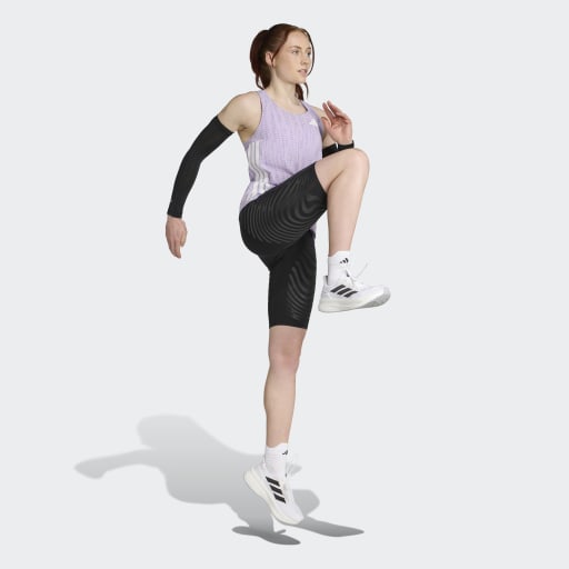 En kvinde i en svedtransporterende Adidas Adizero Singlet, Dame, Powplu/Maglil, sorte kompressionsshorts og hvide Adidas-sneakers løfter sit knæ og sin arm, mens hun laver løbeopvarmning på en ensfarvet baggrund.