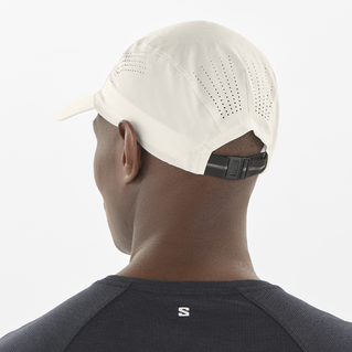 En person ses bagfra iført en Salomon SHKOUT Cap U i Whisper White med ventilationshuller og justerbar pasform, sammen med en mørk skjorte med et lille "S"-logo på den øverste del af ryggen.