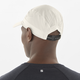 En person ses bagfra iført en Salomon SHKOUT Cap U i Whisper White med ventilationshuller og justerbar pasform, sammen med en mørk skjorte med et lille "S"-logo på den øverste del af ryggen.