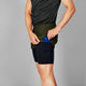 En person i Saysky 2 in 1 Pace Shorts 5'', Herre, Green og en sort t-shirt lægger en blå smartphone i den indvendige sidelomme. Baggrunden er almindelig grå.