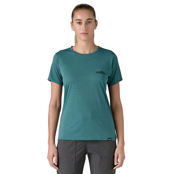 En kvinde står med ansigtet fremad i en Patagonia W's Cap Cool Daily Graphic Shirt, Dame, 73 Skyline/Wetland Blue X-Dye med UPF-beskyttelse og mørkegrå bukser på en almindelig hvid baggrund.