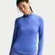En kvinde viser Nike Swift UV Half Zip Running Top, Dame i safir/reflekterende sølv, med lange ærmer og en kvart lynlås, stående mod en almindelig baggrund med et neutralt udtryk.