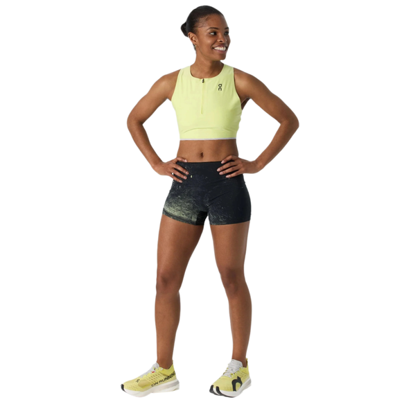En kvinde står med hænderne på hofterne og smiler i en lysegul ærmeløs crop top og On Race Tights Short, Dame, Black/Limelight. Hun har lysegule løbesko på mod en hvid baggrund.