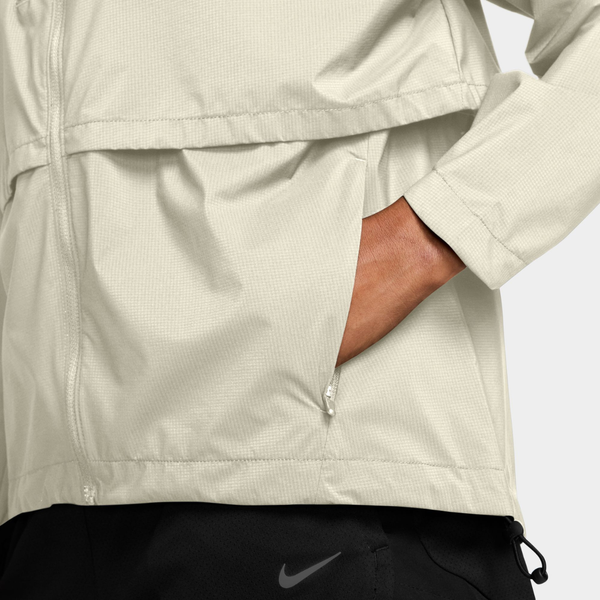 Nærbillede af en lysbeige Nike Storm-FIT løbejakke til kvinder med lynlås i farvekombinationen Sail/Reflective Silv, hvor der ses en hånd i en lomme.