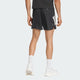 En person står med ansigtet vendt mod kameraet iført sorte Adidas Adi365 Shorts, Herre, en sort skjorte, hvide strømper og hvide sneakers. Den ensfarvede lyse baggrund fremhæver dette løb og fitness-look.