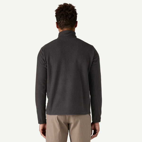 En person vender ansigtet væk fra kameraet og er iført en Patagonia R1 Air Zip Neck, Herre, Black fleece og lysebrune bukser på en almindelig lys baggrund.
