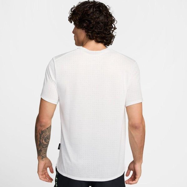 En mand med krøllet mørkt hår og en tatovering på venstre underarm står med ryggen til kameraet iført en Nike Miler Short-Sleeve Running Top, Herre, Summit White/Reflective Silver - en ideel løbetrøje herre - sammen med sorte shorts på en ensfarvet baggrund.