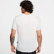 En mand med krøllet mørkt hår og en tatovering på venstre underarm står med ryggen til kameraet iført en Nike Miler Short-Sleeve Running Top, Herre, Summit White/Reflective Silver - en ideel løbetrøje herre - sammen med sorte shorts på en ensfarvet baggrund.