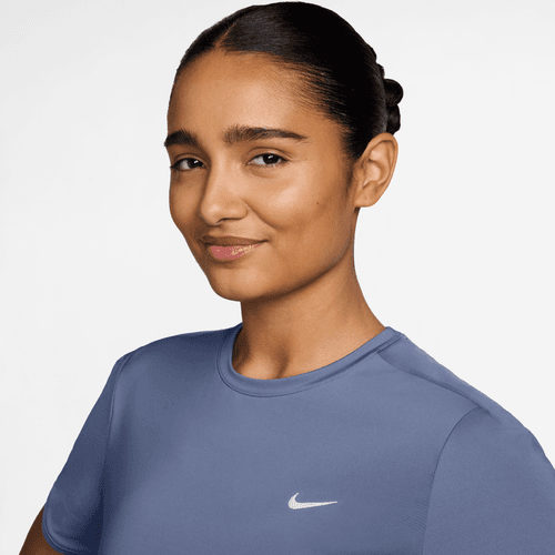 En kvinde med brun hud og mørkt hår, der er trukket tilbage, bærer en lyslilla Nike Dri-FIT kortærmet løbetop (World Indigo/Reflective Silver) med et hvidt logo. Hun vender sig mod kameraet og smiler let mod en almindelig hvid baggrund.