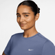 En kvinde med brun hud og mørkt hår, der er trukket tilbage, bærer en lyslilla Nike Dri-FIT kortærmet løbetop (World Indigo/Reflective Silver) med et hvidt logo. Hun vender sig mod kameraet og smiler let mod en almindelig hvid baggrund.