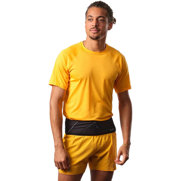 En person i gul kortærmet skjorte og shorts står med et neutralt udtryk og bærer Salomon Pulse Løbebælte, Unisex, Black om livet. Baggrunden er almindelig hvid.