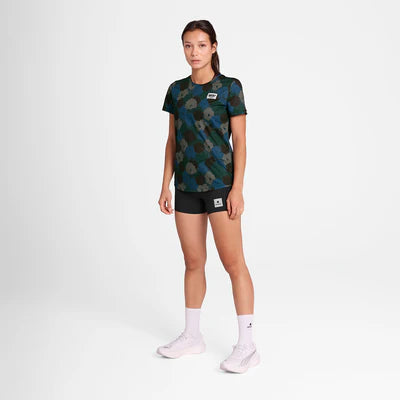 En kvinde kigger fremad mod en hvid baggrund iført Saysky W Flower Combat T-Shirt, Dame, Blue Aop med sorte shorts, hvide strømper og hvide sneakers. Hun har et neutralt udtryk.