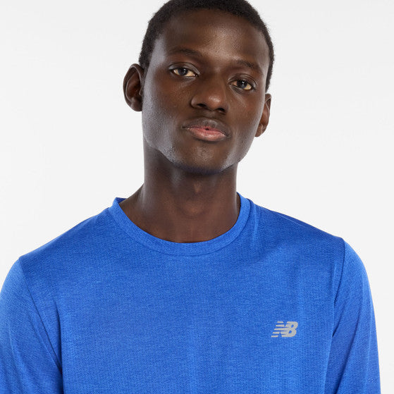 En mand i New Balance Athletics Long Sleeve, Herre, Blue Gemstone Heather, kigger på kameraet med et neutralt udtryk mod en almindelig hvid baggrund.