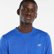 En mand i New Balance Athletics Long Sleeve, Herre, Blue Gemstone Heather, kigger på kameraet med et neutralt udtryk mod en almindelig hvid baggrund.