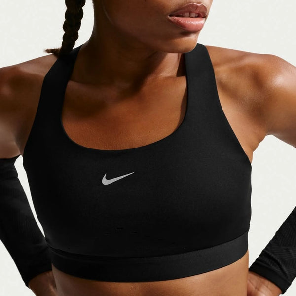 En kvinde bærer Nike Swift Hs Bra, Dame i sort/sølv med sorte ærmer og hænderne på hofterne. Kun hendes overkrop og en del af hendes ansigt vises mod en almindelig lys baggrund.