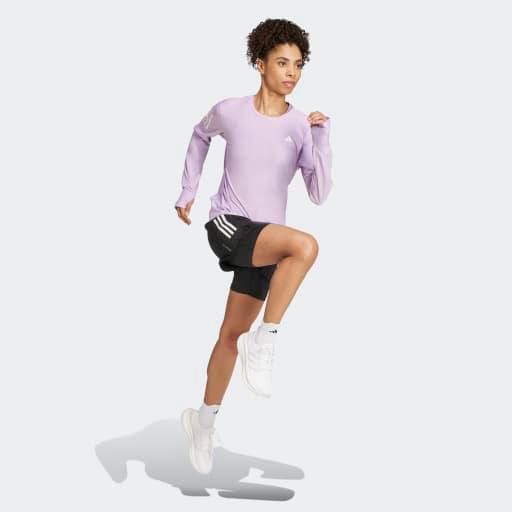 En kvinde i lyslilla Adidas Otr B Ls løbetrøje Dame Powplu, sorte shorts og hvide sneakers er midt i et skridt mod en almindelig lysegrå baggrund og indtager en løbe- eller træningspositur.