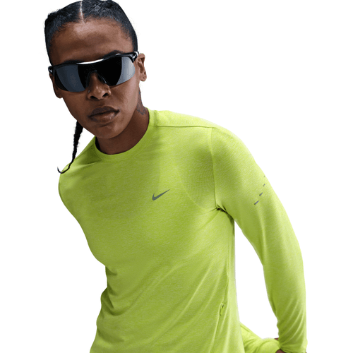 En kvinde med flettet hår bærer sorte solbriller og en neongulgrøn Nike Dri-FIT UV Long-Sleeve Crew-Neck Running Top, Cyber/Htr/Reflective Silv, og ser alvorlig ud mod en hvid baggrund. Toppen giver UV-beskyttelse til udendørs løbeture.