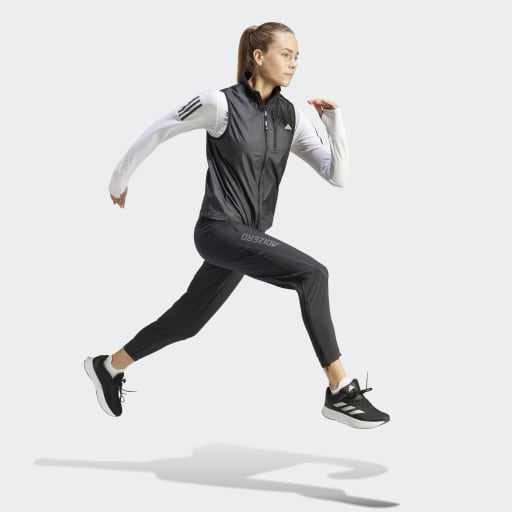 En kvinde i dame løbetøj, herunder Adidas Otr B Vest i sort, joggers og sneakers, er fanget midt i luften i et løbespring mod en almindelig lys baggrund.