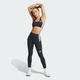 En kvinde i sort Adidas Adizero Medium Support Run Bra (Dame) og leggings med hvidstribede detaljer strækker sin venstre arm ud over hovedet, parret med hvide sneakers med flerfarvede detaljer - ideelt til løb.