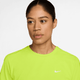 En kvinde med mørkt hår, der er bundet tilbage, iført en Nike Dri-FIT kortærmet løbetop til kvinder i Cyber/Reflective Silver, kigger lidt til siden mod en almindelig hvid baggrund.