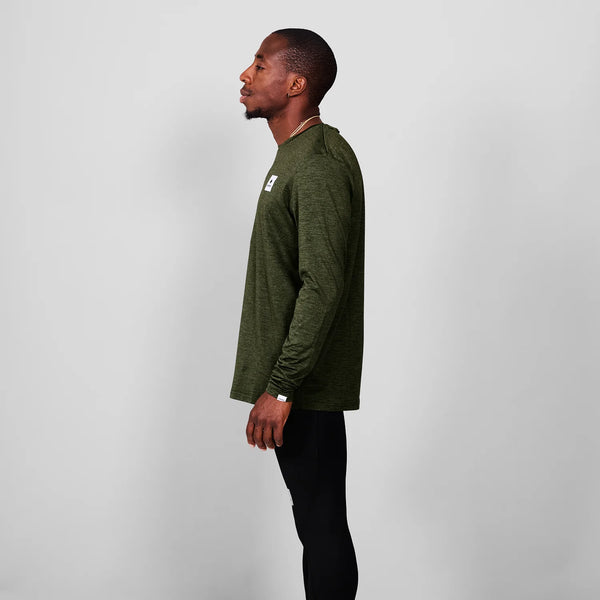En mand står i profil mod venstre, iført en Saysky Clean Pace Long Sleeve, Herre, Green med sorte bukser på en almindelig lysegrå baggrund.