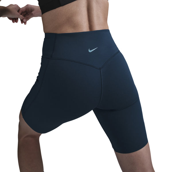Personen strækker sig i Nike Medium-Support High-Waisted 8" Biker Shorts til kvinder, Black/Anthracite/Dk Smoke Grey, og en sort top, her set bagfra mod en almindelig hvid baggrund.