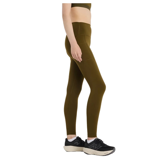 En person iført New Balance's Sleek Pocket High Rise Legging i Woodland, parret med en olivenfarvet sports-bh og sorte og hvide sportssko, står sidelæns mod en almindelig hvid baggrund.