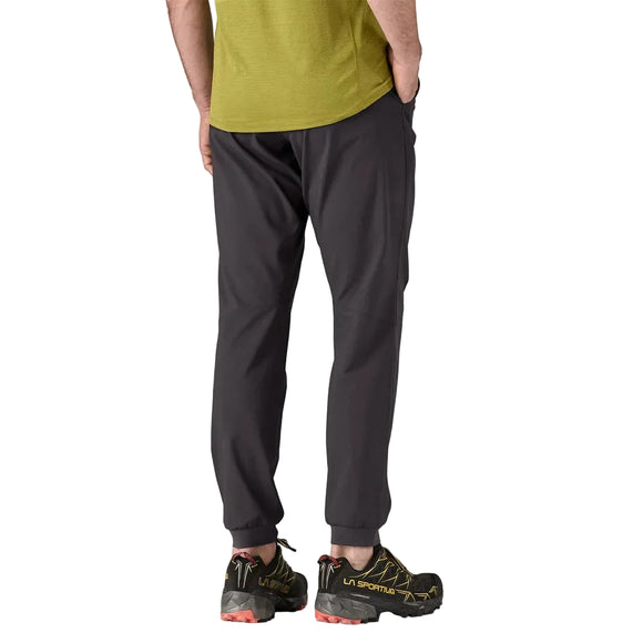 En person ses bagfra iført en grøn T-shirt, Patagonia Terrebonne Joggers til mænd i sort og sorte trail-løbesko med gule detaljer, stående på en almindelig lys baggrund.