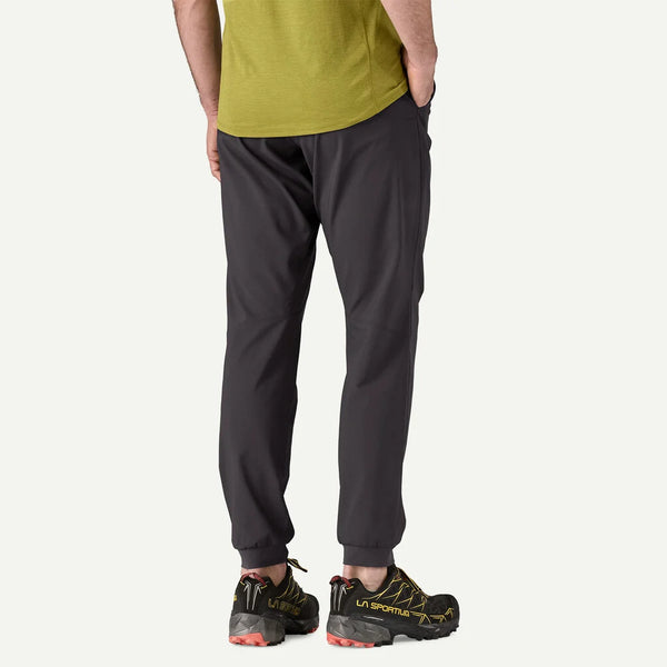 En person ses bagfra iført en grøn T-shirt, Patagonia Terrebonne Joggers til mænd i sort og sorte trail-løbesko med gule detaljer, stående på en almindelig lys baggrund.