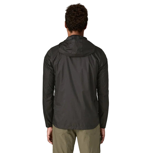 En person står med ryggen til kameraet på en almindelig, lys baggrund iført Patagonias Houdini Jacket, Herre i Black m/Black og olivengrønne bukser.