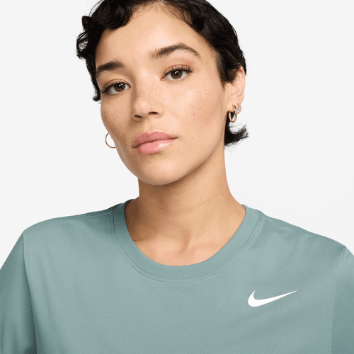 En person med kort, mørkt hår og guldøreringe bærer en lys blågrøn Nike Dri-FIT Dame Cannon T-shirt med et hvidt logo og står på en almindelig lys baggrund.
